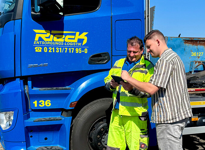 Zwei Personen stehen neben einem Lkw-Führerhaus, das mit Rieck gebrandet ist, und schauen gemeinsam auf einen Handheld.